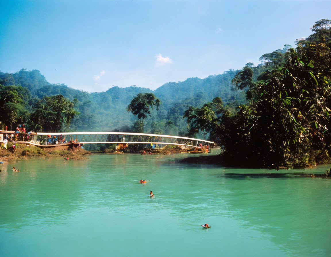 Scenic turquoise river with swimmers, white bridge, lush green forest, and blue sky