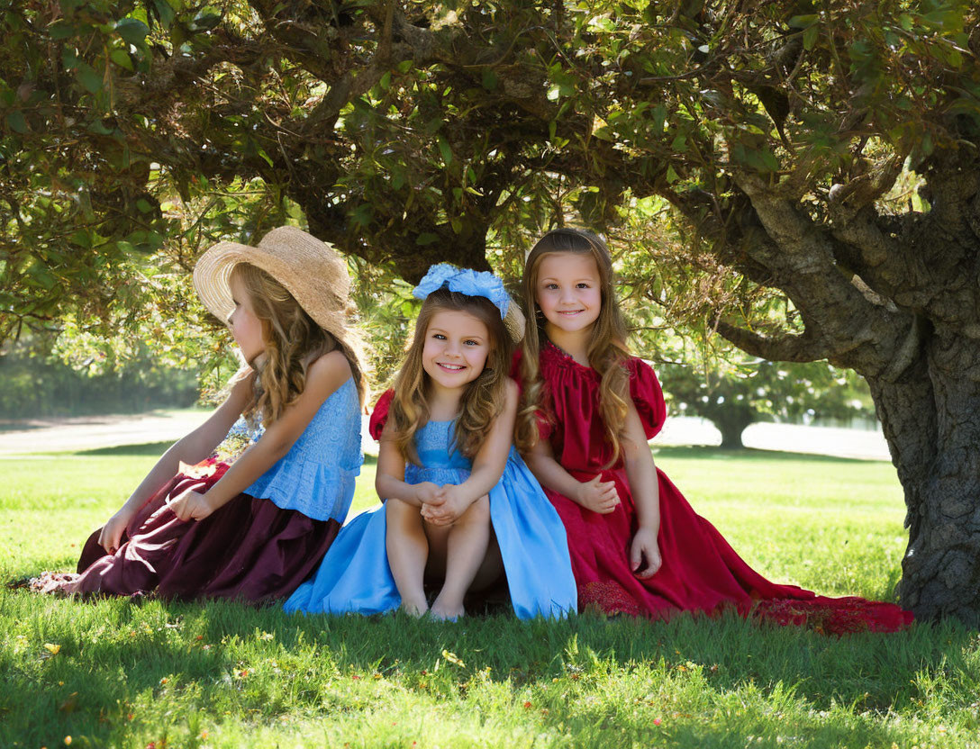 Three girls in colorful dresses under a tree on a sunny day, one in a straw hat