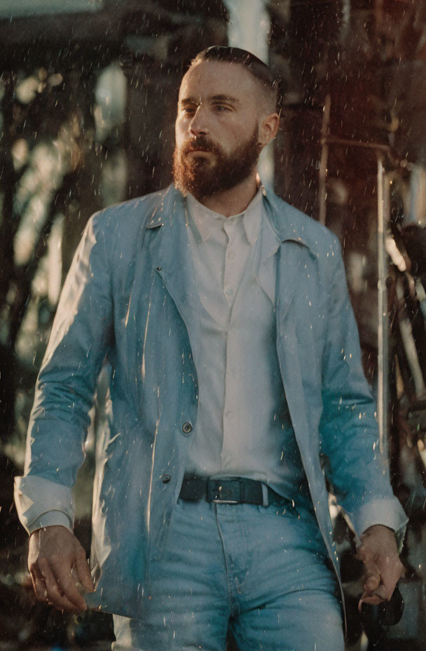 Bearded man in light blue suit standing in rain with pensive expression