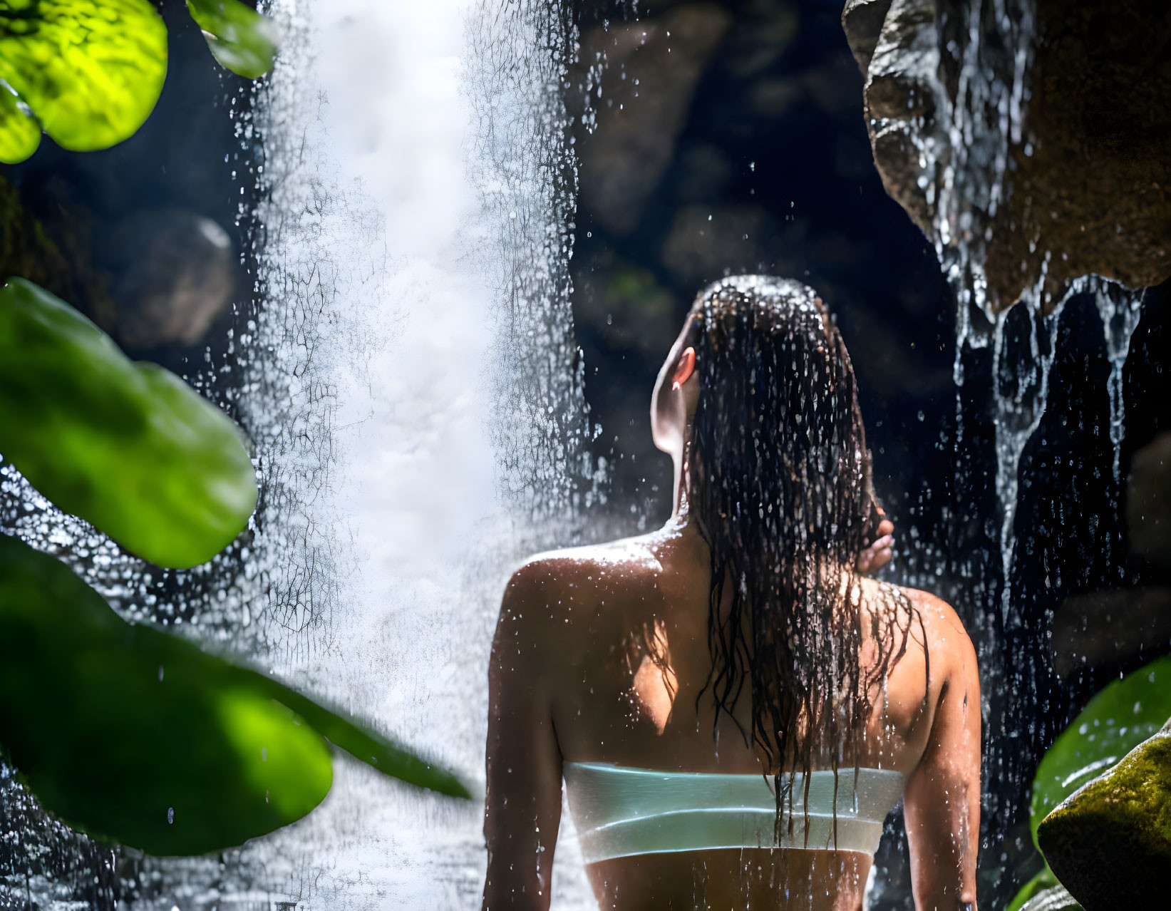 Person standing under waterfall with green foliage and sunlight filtering through droplets.