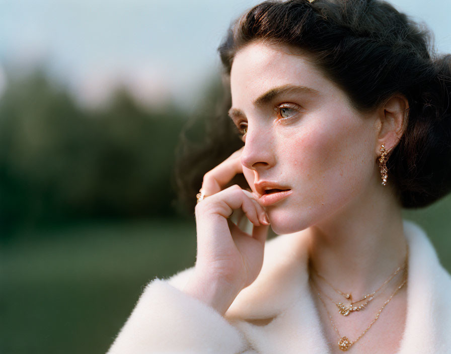 Dark-haired woman in updo with earrings and necklace gazing into distance against natural backdrop