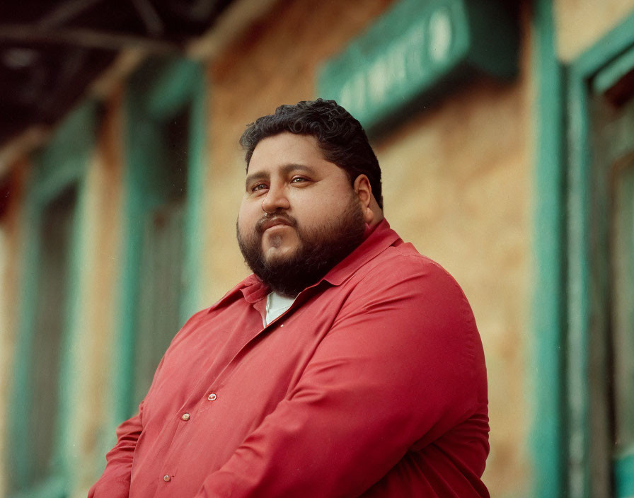 Bearded man in red shirt with thoughtful expression leaning against wall with windows.