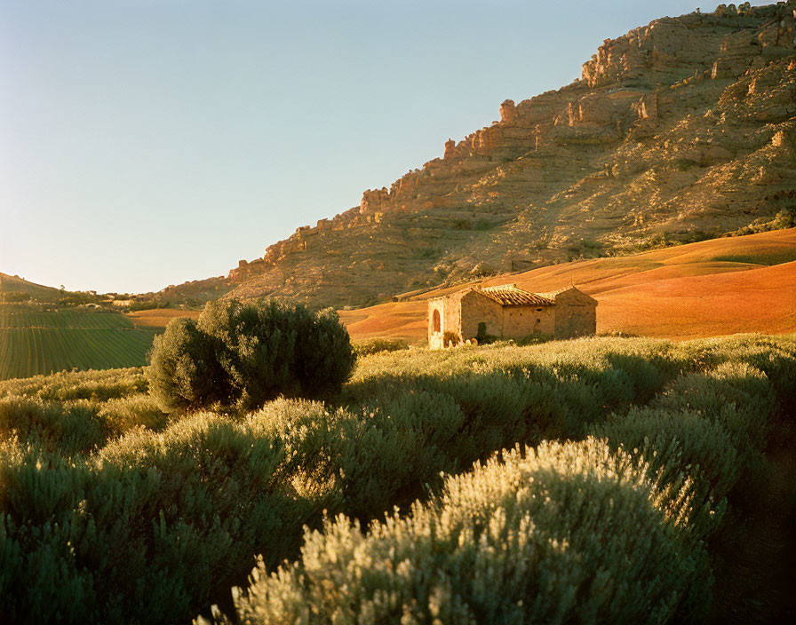 Rustic stone building in lush fields with rocky cliffs under warm sunlight