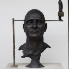 Sculpture of human head with crossbar and miniature figures on plain background