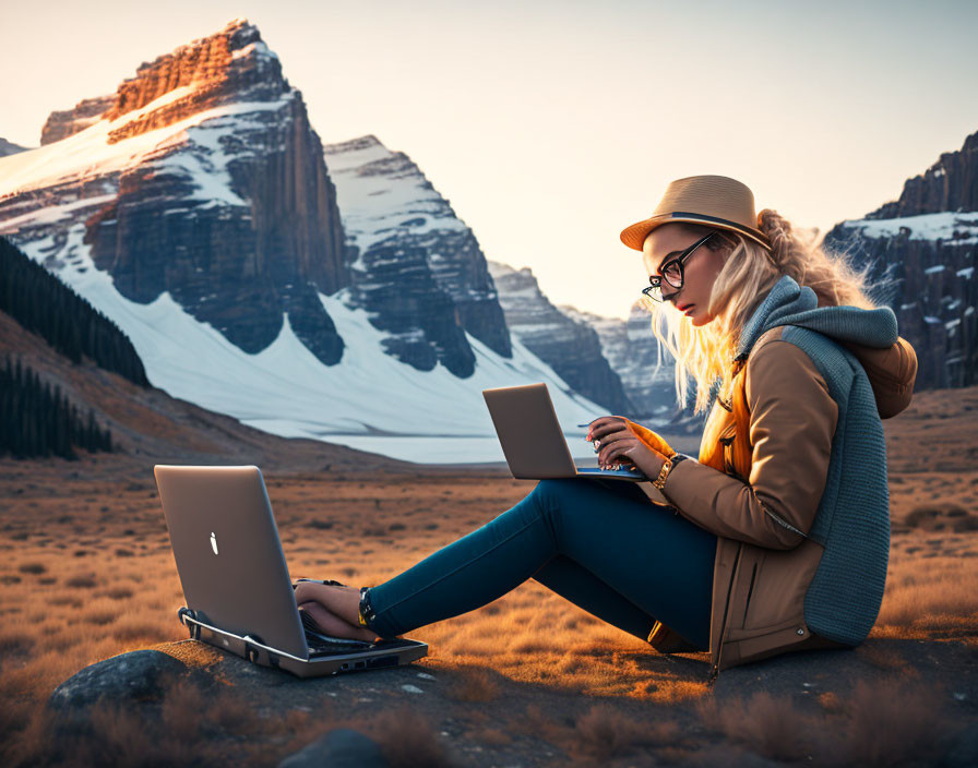 Woman in glasses and hat uses laptop with snowy mountains at sunset