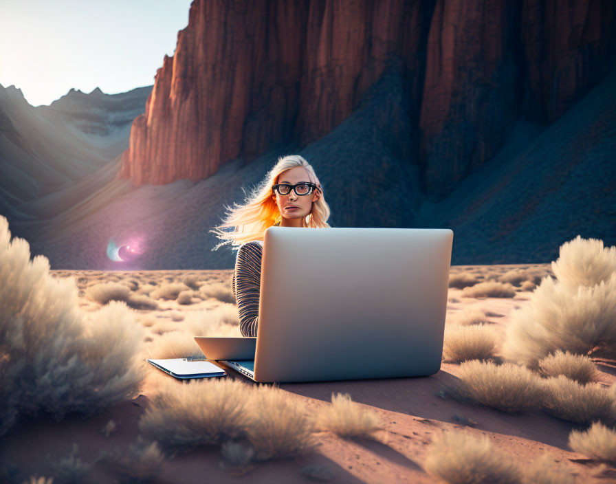 Woman with glasses at laptop in desert with red cliffs and evening glow