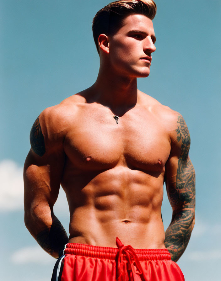 Muscular Shirtless Man with Tattoos in Red Shorts and Pendant against Blue Sky