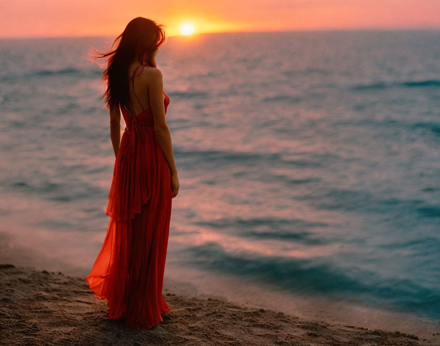 Woman in red dress on beach at sunset watching the sun setting over the sea