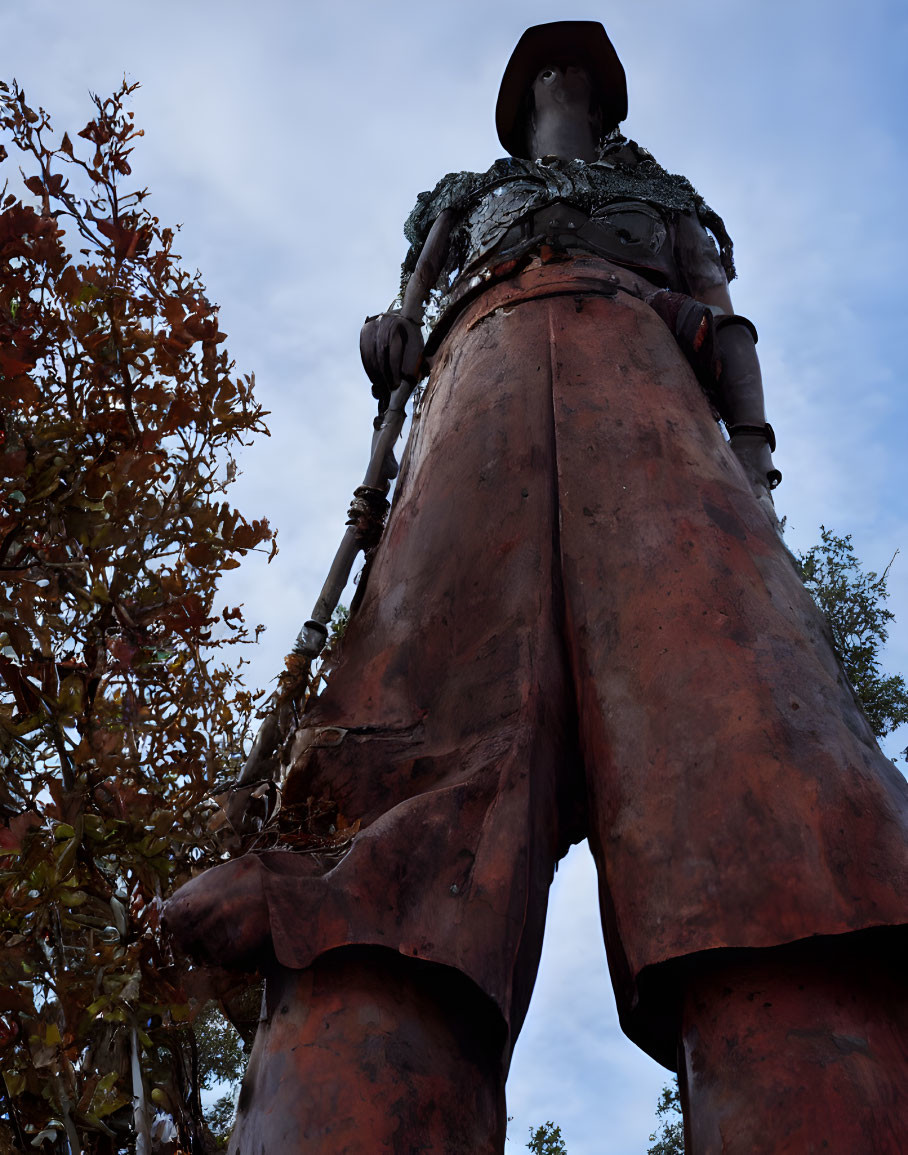 Metal statue of figure with hat among trees against blue sky.