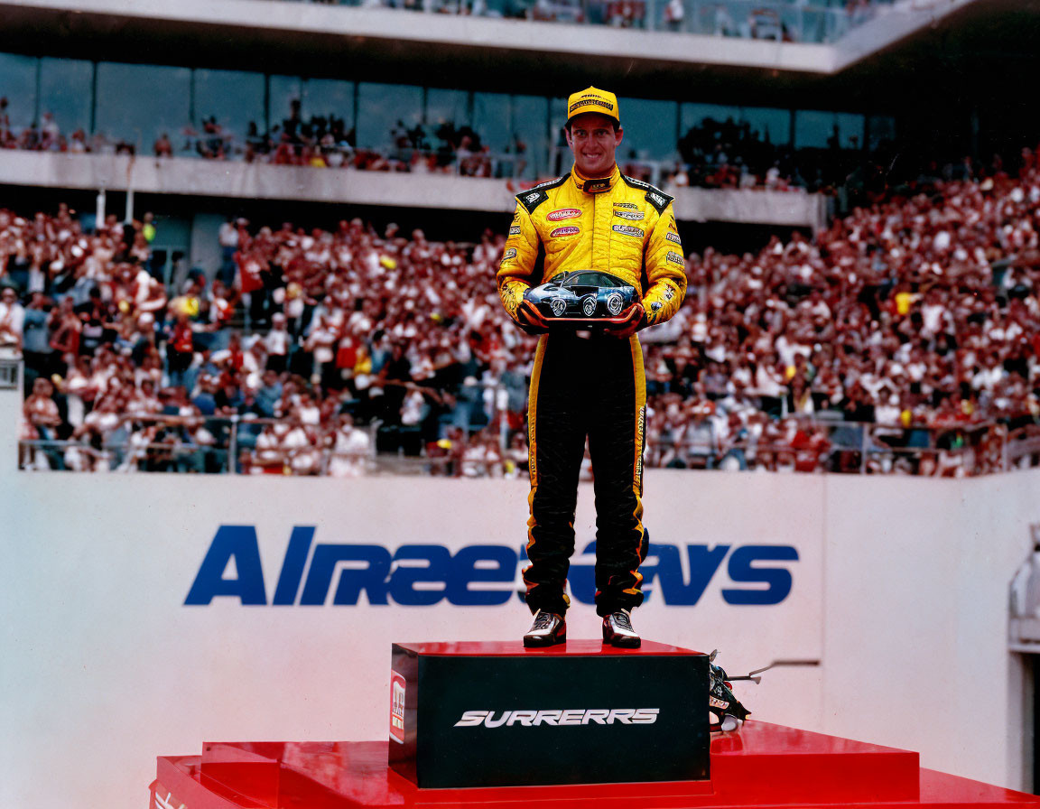 Smiling racing driver in yellow suit on winner's podium with crowd and sponsor logos