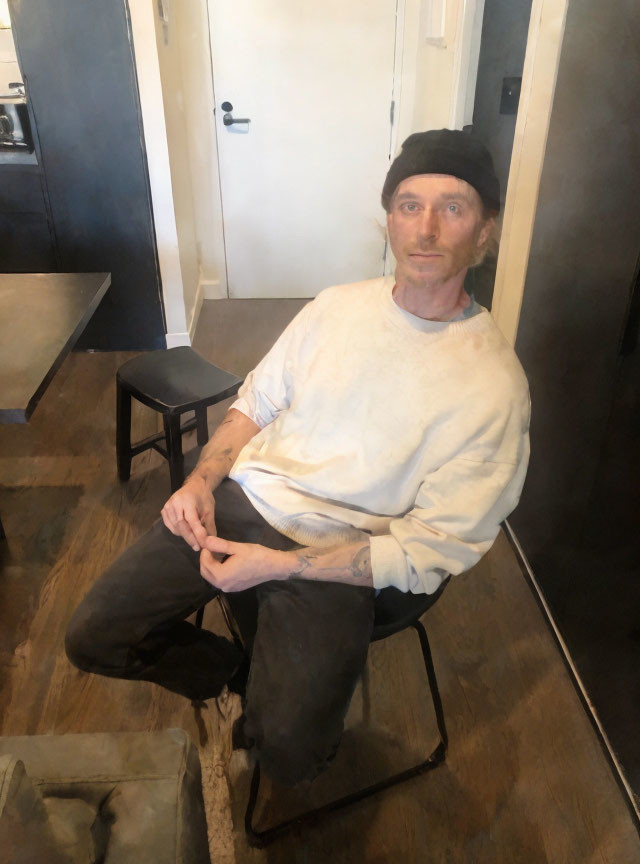 Man in white shirt and black hat sitting on folding chair in room with closed door and black stool.