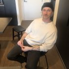 Man in white shirt and black hat sitting on folding chair in room with closed door and black stool.