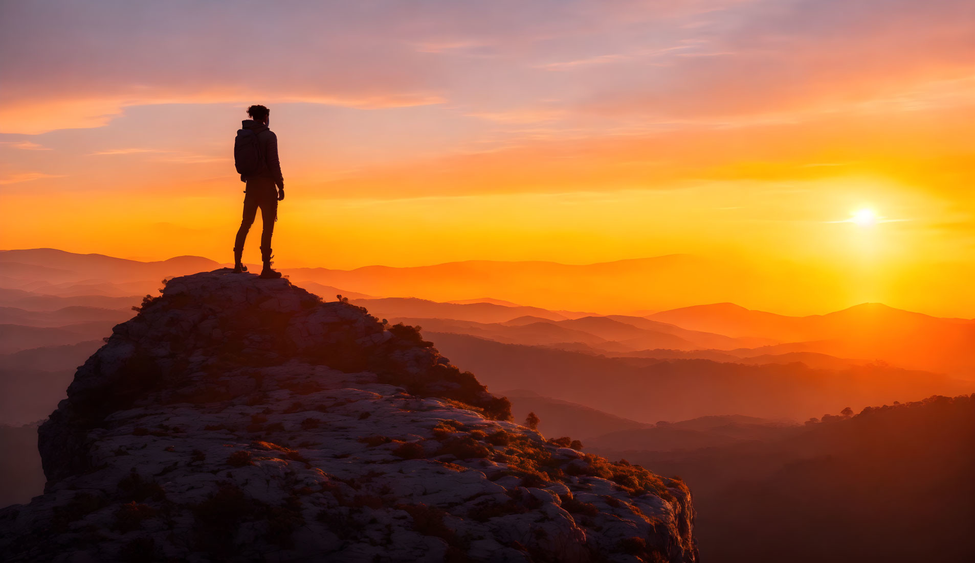 Person standing on mountain peak at vivid sunset with layered hills.