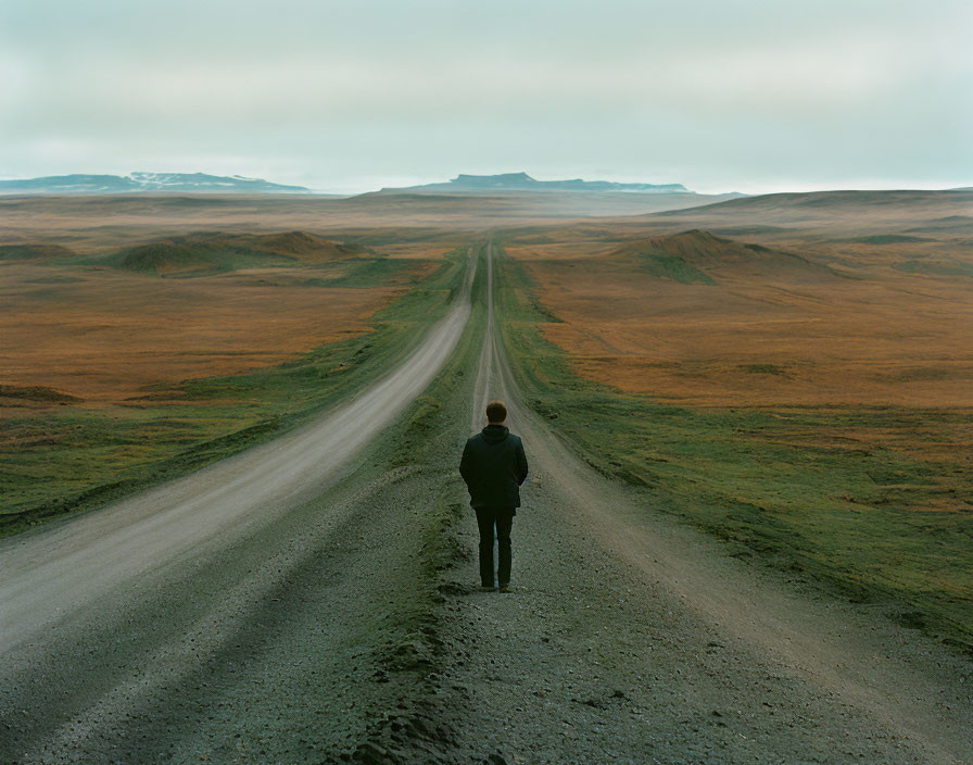 Person standing on winding dirt road amidst grass-covered hills under cloudy sky