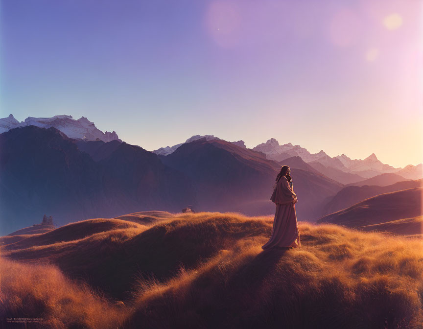 Solitary figure on grassy hill gazes at majestic mountain range at sunset