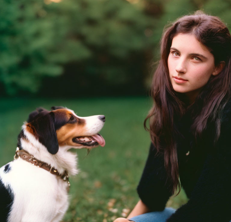 Young woman with long hair kneeling beside tricolor dog in grassy area