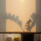 Natural sunlight through sheer curtains creates shadow on wall with coffee cup and potted plant.