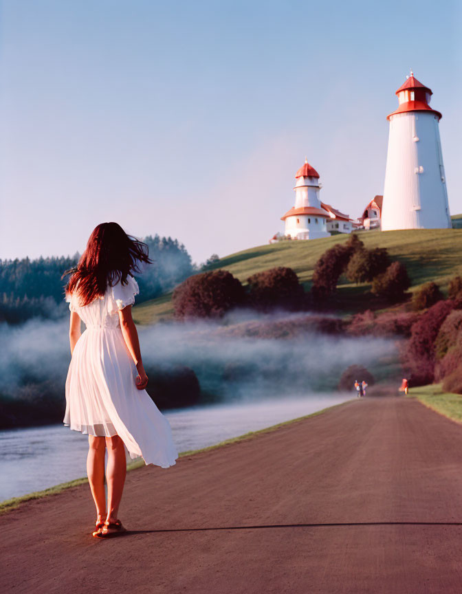 Woman in white dress walking to riverside lighthouse under clear blue sky