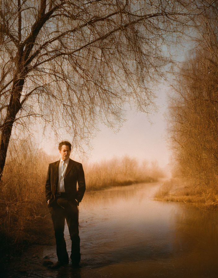 Businessman in suit standing by river under tree canopy with mist.