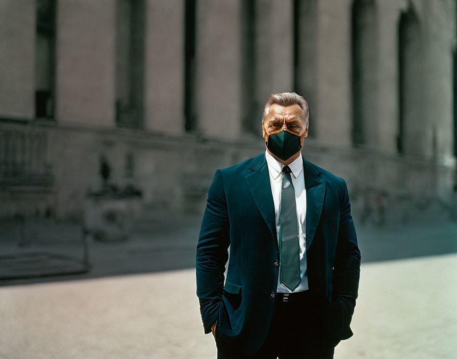 Man in dark suit and tie with black mask in front of blurred classical building.