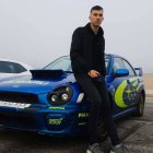 Man in navy jacket leans on classic sports car with vintage car in background on open road