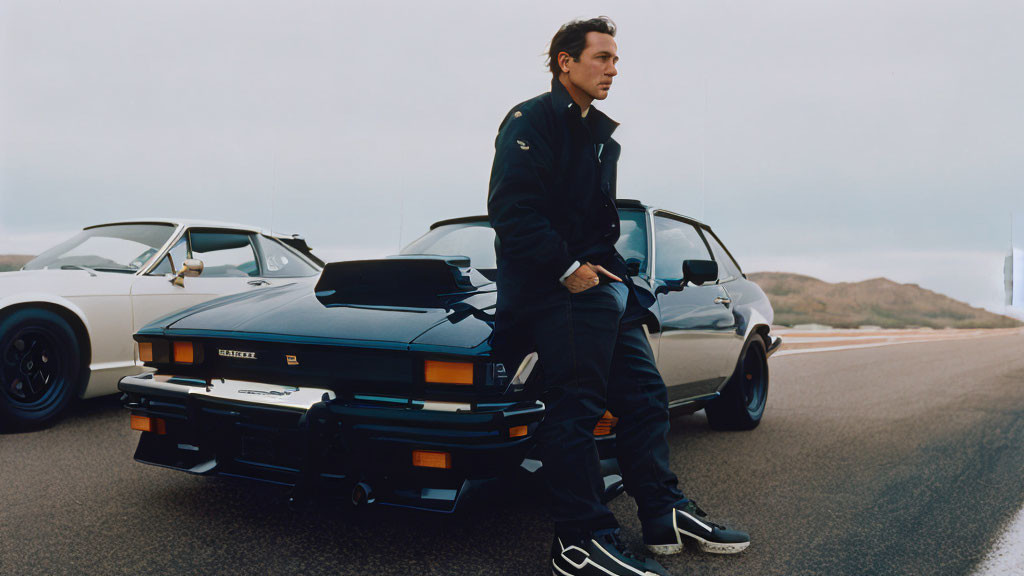 Man in navy jacket leans on classic sports car with vintage car in background on open road