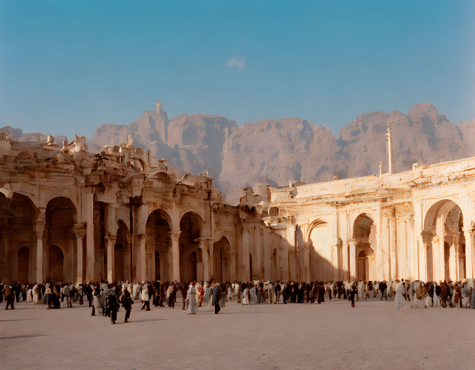 Crowd gathers at ancient ruins with towering cliffs and clear sky