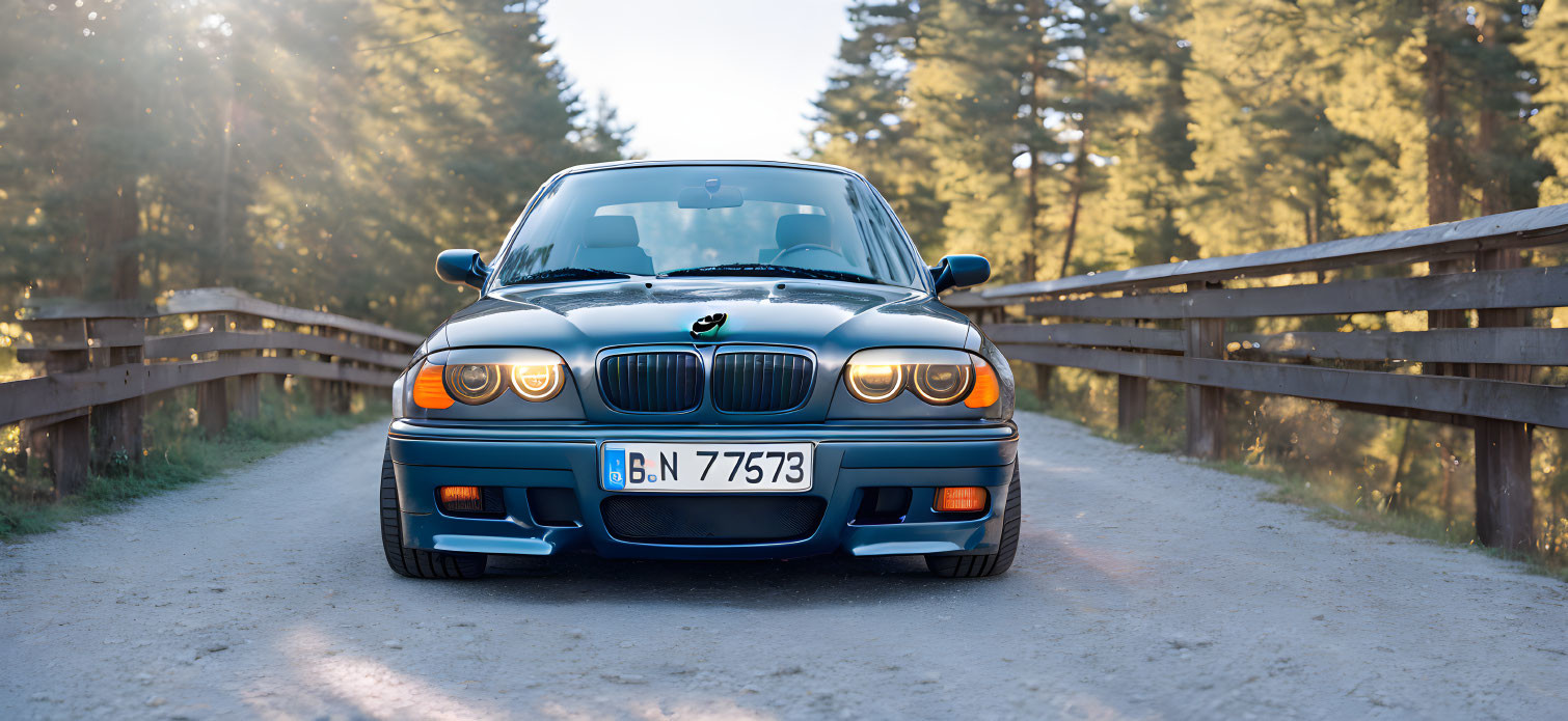 Vintage BMW car parked on countryside road with wooden fence, trees, and sunlight glare.