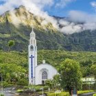 White Church with Spire in Greenery and Mountains Under Dramatic Sky