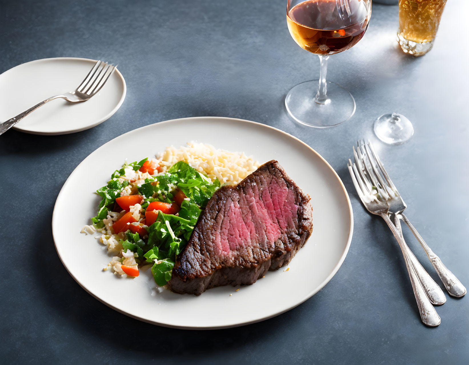 Plate of medium-rare steak with rice, tomatoes, parsley, wine, silverware on dark table