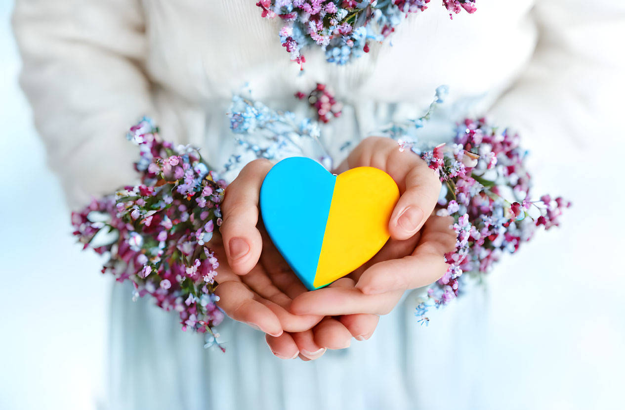 Multicolored heart paper cutout held by hands amidst purple flowers