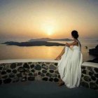 Woman sitting on rocky shore at sunset with tranquil sea and distant mountains.