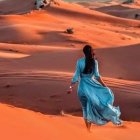 Person in flowing blue dress and wide-brimmed hat in orange sand dunes at sunset