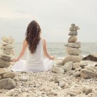 Woman in white dress gazes at sea by shore with rock cairn and colorful flowers.