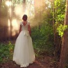 Woman in white gown in sunlit autumn forest with tall trees and fallen leaves