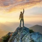 Person standing triumphantly on cliff at sunset with misty hills and mountains.