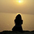 Woman sitting on calm beach at sunset with serene ocean and orange sky