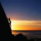 Person's silhouette on cliff at sunset with sun, clouds, mountains.