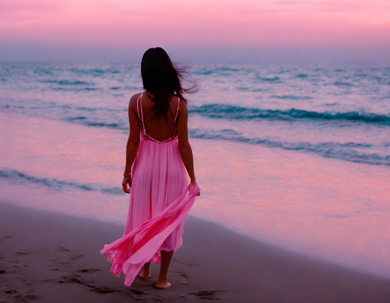 Woman in Pink Dress Walking on Beach at Sunset with Waves and Pink-Blue Sky