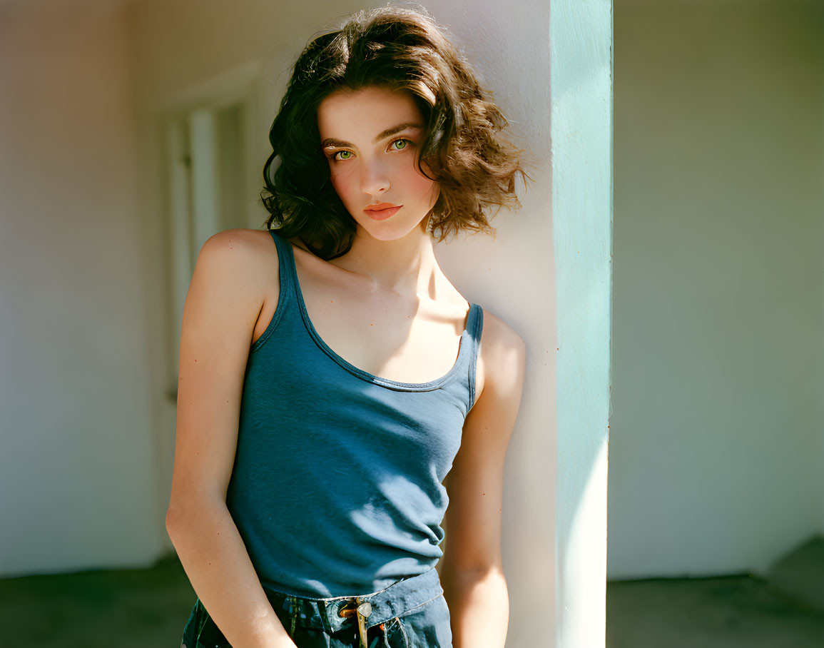 Dark-haired woman in blue tank top against white wall in soft sunlight