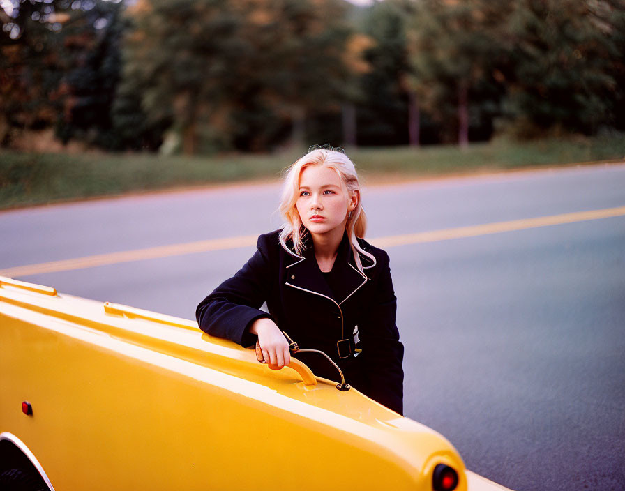 Person leaning on yellow vehicle in dark coat, looking pensive with blurred background