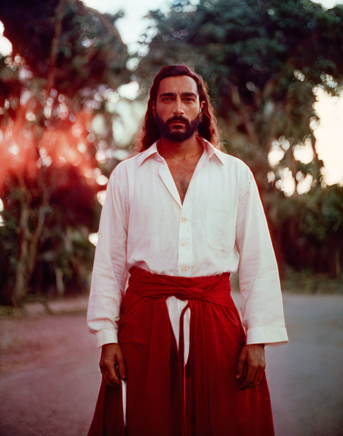 Bearded man in white shirt and red sash confidently standing on foliage-filled path