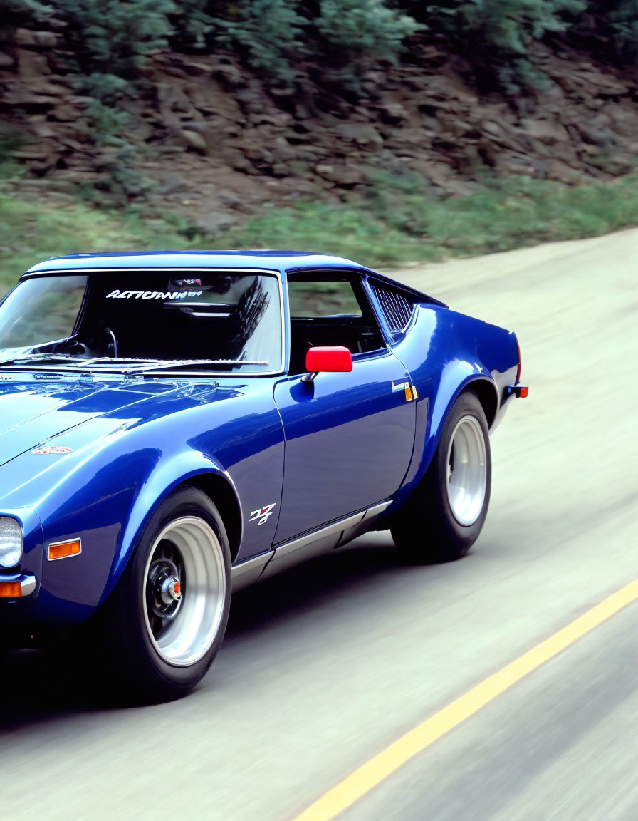 Blue vintage sports car with white stripes and "Battmobile" text driving on a road amidst green