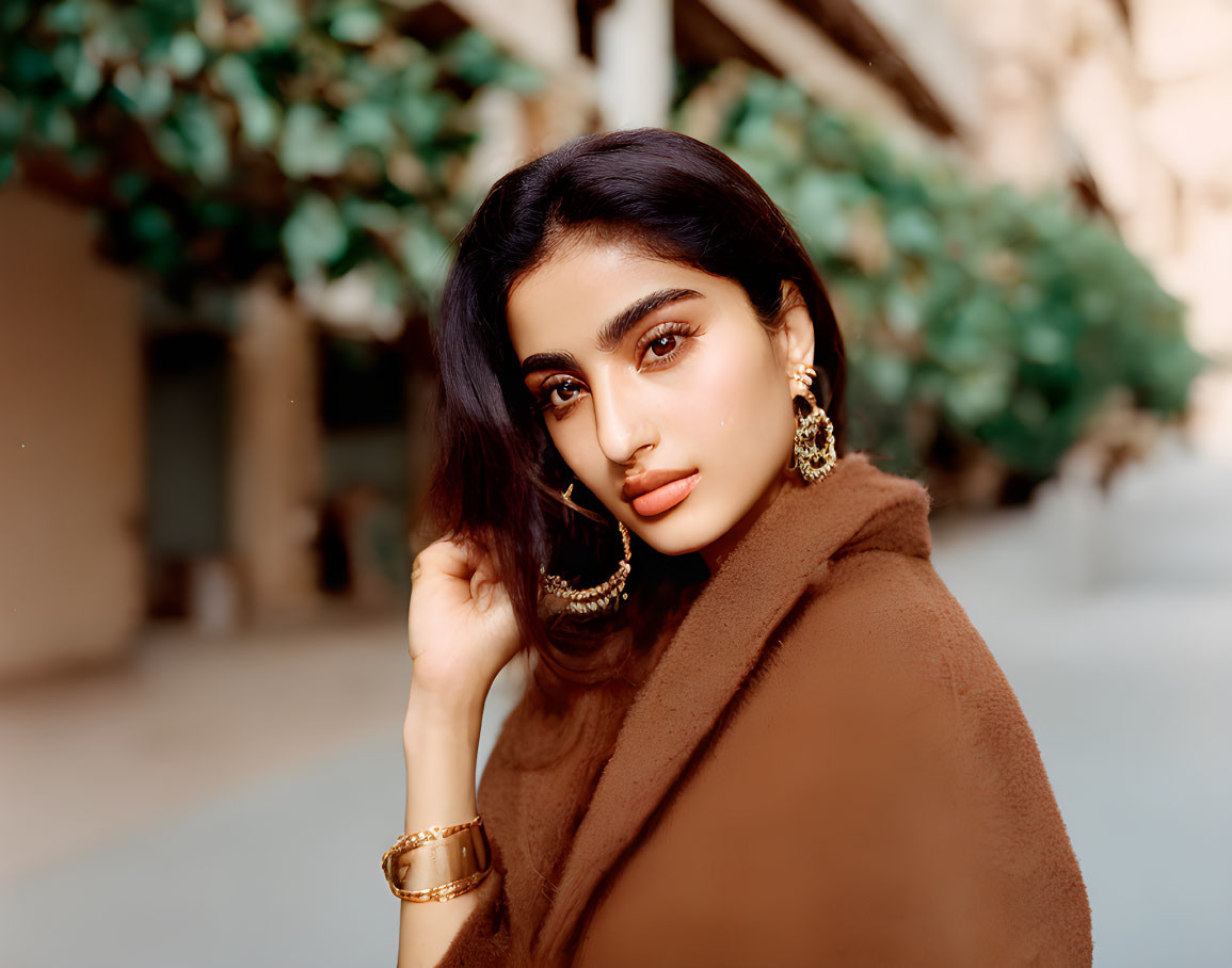 Dark-haired woman in gold jewelry wrapped in brown cloak against green foliage