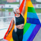 Smiling person with white hair holding rainbow flag near waterfall