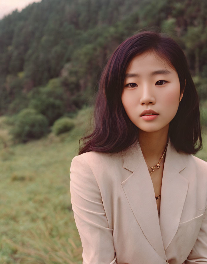 Dark-haired woman in beige blazer amidst trees in field.