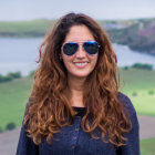 Woman with Curly Hair in Sunglasses and Blue Leather Jacket Against Serene Landscape