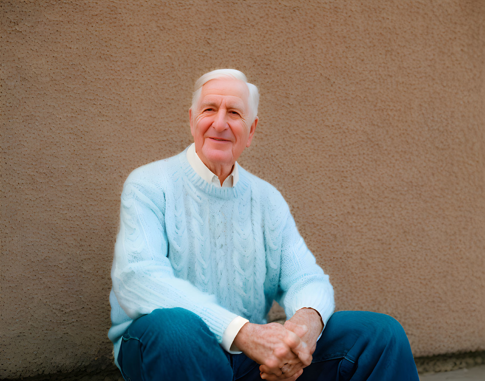 Elderly man in light blue sweater and jeans sitting against beige wall