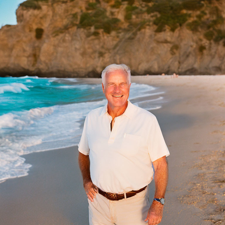 Smiling man in white attire on sandy beach with cliffs and waves