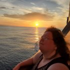 Girl on boat watching dolphin leap at sunset over ocean with mountains.